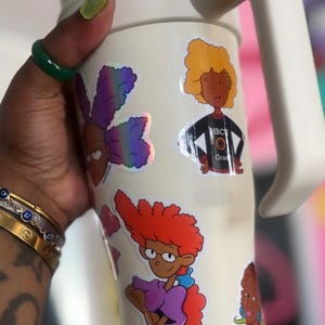 May include: White travel mug with a handle, featuring colorful cartoon stickers of characters with afro hairstyles. The stickers include the text "HBCU Grad".
