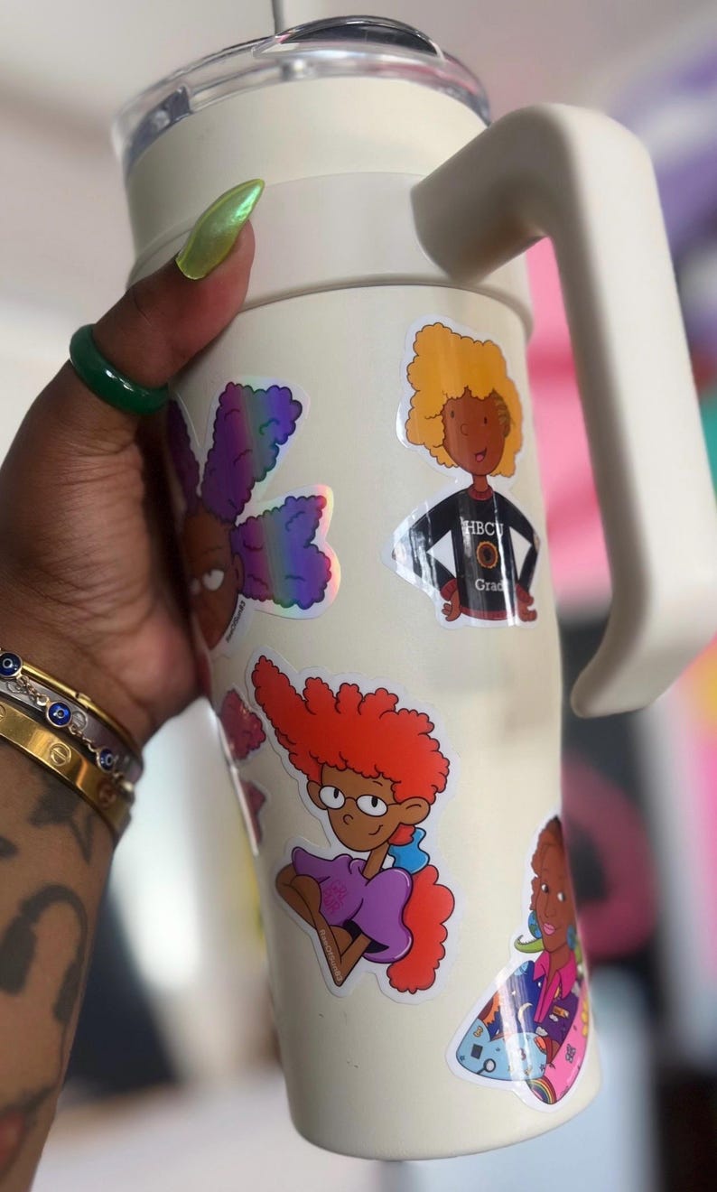 May include: White travel mug with a handle, featuring colorful cartoon stickers of characters with afro hairstyles. The stickers include the text "HBCU Grad".