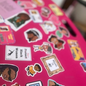 May include: A pink surface covered with colorful stickers featuring cartoon characters, illustrations, and text. Some of the stickers include the words "Protect Black Women", "Library Card", and "Read is Art".