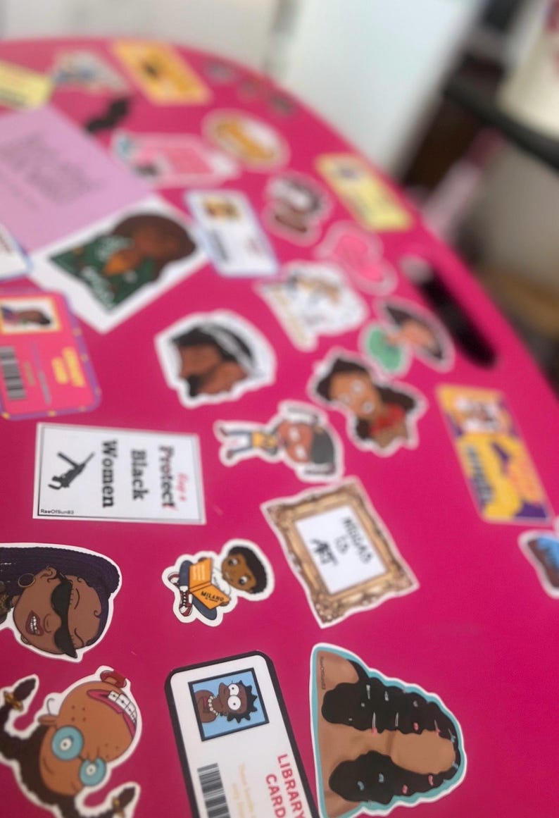 May include: A pink surface covered with colorful stickers featuring cartoon characters, illustrations, and text. Some of the stickers include the words "Protect Black Women", "Library Card", and "Read is Art".