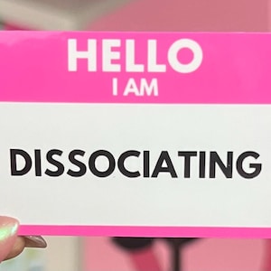 May include: A pink name tag with the words "HELLO I AM" at the top and "DISSOCIATING" in bold black letters below. The name tag is rectangular and held by a person.