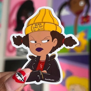 May include: A die-cut sticker featuring a cartoon character with dark brown pigtails, wearing a yellow beanie with the text BLACK GIRLS GOT THE JUICE, a black leather-style jacket over a red shirt, and dark purple lipstick with a stern expression.
