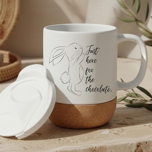 Könnte beinhalten: Weiße Keramik-Tasse mit Korkboden und weißem Deckel. Die Tasse zeigt eine schwarze Strichzeichnung eines Kaninchens und den Text "Just here for the chocolate."