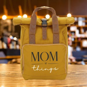 Op de afbeelding: Mosterdgele rugzak met bruine bandjes en een zwarte gespsluiting. De voorzak heeft de tekst "MOM things" in zwart en wit. De tas heeft een handvat aan de bovenkant en een rol-top ontwerp.