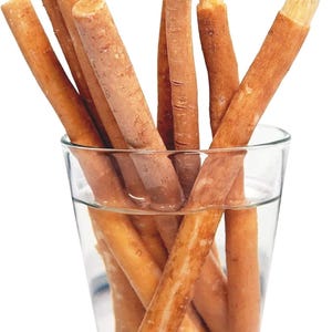 Könnte beinhalten: Ein klares Glas gefüllt mit Wasser und mehreren braunen Miswak-Sticks. Die Enden einiger Stöcke sind ausgefranst und ähneln einer Bürste. Die Stöcke sind zylindrisch und variieren in der Länge, mit einer hellbraunen Farbe.