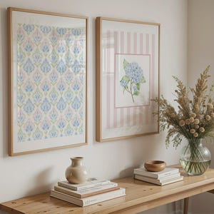 Könnte beinhalten: Zwei gerahmte Wandbilder. Eines zeigt ein blaues, rosa und grünes Blumenmuster. Das andere hat einen rosa gestreiften Hintergrund mit einer blauen Hortensien-Illustration. Ein Holztisch mit Büchern, einer Vase und einer Schale.