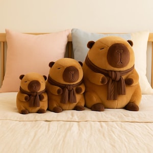 Può includere: Tre orsacchiotti di peluche di diverse dimensioni, ognuno con una sciarpa marrone. Gli orsi hanno gli occhi chiusi e un colore marrone chiaro, con accenti marroni scuri sul muso, sulle orecchie e sulle zampe. Sono disposti su un letto.