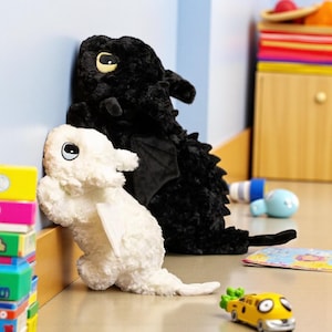 May include: A black plush dragon toy with large yellow eyes and a white plush dragon toy with large blue eyes, both featuring small wings and textured fur, positioned against a light blue wall next to a stack of colorful blocks and a yellow toy car.