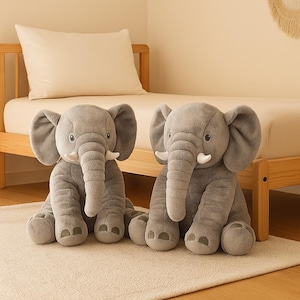 Può includere: Due elefanti di peluche grigi con grandi orecchie, zanne bianche e proboscide dettagliate. Gli elefanti sono seduti su un tappeto color crema davanti a una struttura letto in legno.
