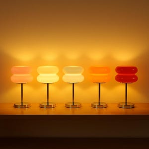 May include: Five table lamps with cylindrical metal bases and rounded, double-tiered glass shades in varying shades of pink, yellow, white, orange, and red. Each lamp emits a warm, soft glow, creating a gradient of light across the wall behind.