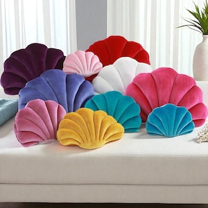 Puede incluir: Una colección de cojines con forma de concha marina en varios colores, incluyendo morado, rosa, rojo, blanco, azul, turquesa y amarillo. Los cojines están colocados sobre un sofá blanco, ofreciendo un acento decorativo y colorido.