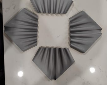 May include: Four gray, abstract, geometric decorative objects arranged in a diamond shape. Each object has a folded, draped appearance, with a textured surface. The objects are placed on a white, marble-like surface.