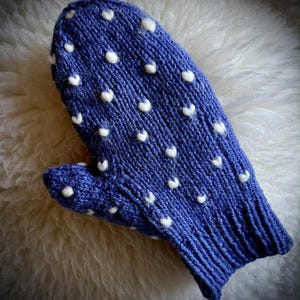 Può includere: Un guanto blu lavorato a maglia con pois bianchi. Il guanto è realizzato con un filato strutturato e ha un polsino a coste. I pois bianchi sono distribuiti uniformemente sulla superficie del guanto.
