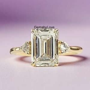 May include: A gold engagement ring featuring an emerald-cut central stone and two smaller side stones. The ring is set against a soft purple and pink backdrop. The text "GemsbyLove" is visible.