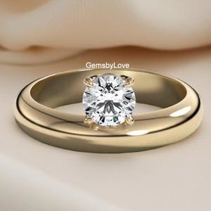 Puede incluir: Un anillo de oro con una gema grande, redonda y transparente. El anillo tiene una banda gruesa y un diseño simple y elegante. La gema está engastada en un engaste de cuatro puntas. El anillo se muestra sobre una tela color crema.