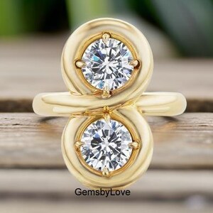 Puede incluir: Un anillo de oro con dos piedras redondas, transparentes y facetadas, engastadas en un marco de metal vertical en forma de ocho. Las piedras están sujetas por cuatro garras cada una. El texto GemsbyLove aparece en letras negras en la base.