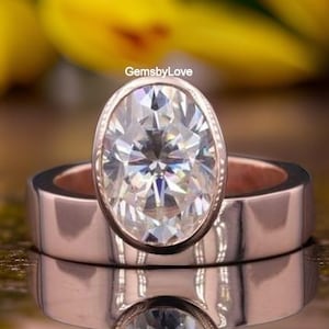 May include: A rose gold ring featuring a large, oval-cut, clear faceted gemstone set in a bezel setting. The band is wide and smooth with a polished finish. The text GemsbyLove is visible above the center stone, which reflects clearly below.