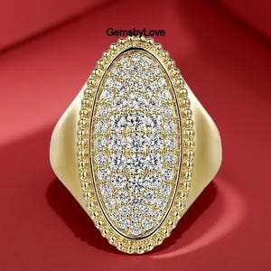 May include: A gold-colored ring with a large, oval-shaped face. The face is covered in small, sparkling white stones, surrounded by a beaded border. The ring has a wide band and is set against a red background.