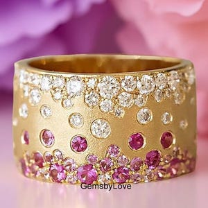 May include: A wide gold band ring featuring a textured matte finish, embellished with a gradient of flush-set round stones. The design transitions from clear white stones at the top to vibrant pink stones concentrated at the bottom edge.