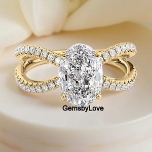 May include: A gold engagement ring with a large oval-cut diamond center stone. The band features a split shank design with pavé-set diamonds. The ring is displayed on a white surface with a soft, cream-colored background. The text "GemsbyLove" is visible.
