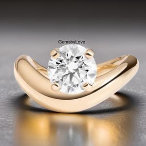 Puede incluir: Un anillo de oro con un gran diamante redondo y transparente en el centro. La banda tiene un diseño curvo que abraza el diamante. El anillo se muestra sobre un fondo neutro.