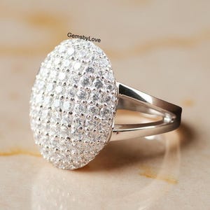 Puede incluir: Un anillo de plata con una gran cara ovalada cubierta de pequeñas piedras preciosas transparentes. El anillo tiene un diseño de caña dividida. El fondo es de color claro y neutro.