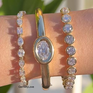 Peut inclure: Trois bracelets en or ornés de pierres précieuses étincelantes. Le bracelet central présente un diamant de taille ovale. Les deux autres bracelets ont une série de diamants ronds et rectangulaires. Le texte "GemsbyLove" est visible.