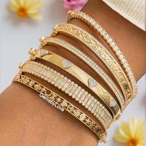 Op de afbeelding: Een stapel van zes goudkleurige armbanden met diverse ontwerpen, waaronder heldere pavé-stenen, baguette-geslepen stenen, hartvormige accenten en gegraveerde symbolen zoals vredestekens, sterren en lieveheersbeestjes. De tekst GemsbyLove is zichtbaar.