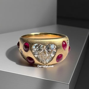 May include: A gold ring with a heart-shaped diamond center stone. The ring is accented with round, red gemstones. The ring has a wide band and a brushed gold finish. The text "GemsbyLove" is visible on the ring.