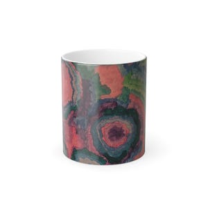 May include: A white ceramic coffee mug with a colorful abstract design. The design features swirling patterns in shades of red, green, and blue. The mug has a standard cylindrical shape and a white interior.