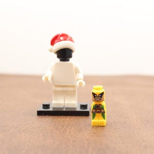 May include: Two Lego figures. One is a white figure with a black head wearing a red and white Santa hat. The other is a yellow figure with a brown head and a green and brown graphic design.