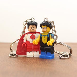 May include: Two Lego-style keychains featuring superhero characters. One figure is red and white with a red cape, the other is yellow, black, and blue. Both have silver key rings attached.