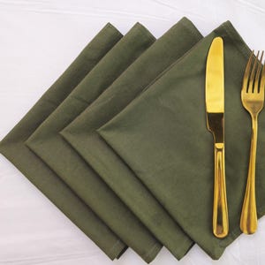 Puede incluir: Vista aérea de cuatro servilletas de tela verde oliva dobladas. Un cuchillo y un tenedor dorados se colocan encima. La cubertería tiene un acabado brillante. Las servilletas están dispuestas en diagonal.