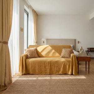 Puede incluir: Un sofá amarillo mostaza con una manta a juego y dos cojines decorativos. La habitación cuenta con cortinas beige, una alfombra de yute y una mesita de noche de madera con un jarrón y libros. La luz del sol crea sombras en el sofá.