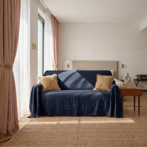 Può includere: Un divano blu navy con una copertura abbinata e due cuscini beige. Il divano è appoggiato su un tappeto di iuta intrecciata. Delle tende rosa incorniciano una finestra a sinistra e un piccolo tavolino di legno a destra.