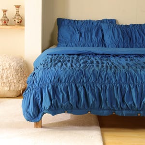 May include: A blue bedspread and pillow set with a gathered, textured design. The bedspread has a ruffled edge and is on a wooden bed frame. The room has a neutral color palette with a fluffy, cream-colored ottoman and decorative vases.