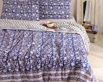 May include: A bed is made with a blue and white floral patterned quilt and matching pillowcases. The quilt has a border with stripes and a floral design. A small wooden tray with a cup of coffee sits on the bed. A mirror and basket are in the background.