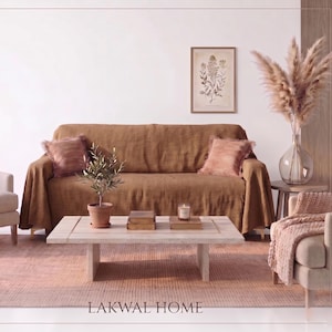 May include: A brown linen sofa cover draped over a couch, featuring two matching brown throw pillows. The scene includes a light-coloured wooden coffee table, a small potted olive tree, a candle, and the text LAKWAL HOME at the bottom.