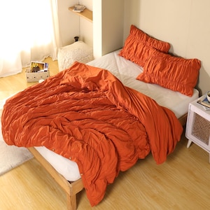 以下が含まれることがあります： 木製のベッドフレームに、オレンジ色のベッドスプレッドと2つのお揃いの枕カバー。寝具は、質感のあるギャザーデザインです。ベッドは白いフィットシーツでできています。部屋はニュートラルなカラーパレットです。