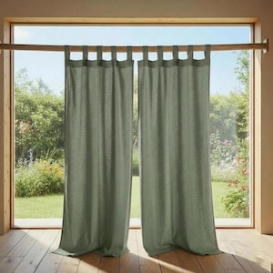 May include: Two sage green linen curtains hanging from a wooden rod in front of a large window. The curtains have tab tops and are slightly textured. The window looks out onto a lush green garden with a wooden floor.