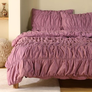 Può includere: Un set di biancheria da letto color malva con un design a balze. Il set include un copripiumino e due federe. La biancheria da letto è realizzata in tessuto morbido ed è su un telaio del letto in legno. Tema: arredamento camera da letto.