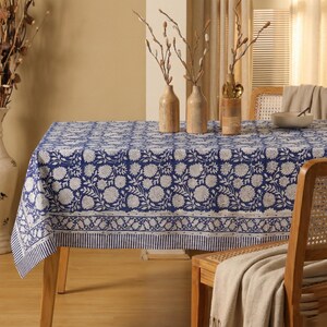 Può includere: Una tovaglia a fantasia floreale blu e bianca copre un tavolo di legno. La tovaglia presenta un motivo floreale ripetuto e un bordo a righe. Vasi decorativi con rami sono sul tavolo e una sedia di legno è in primo piano.