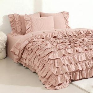 May include: A dusty rose bed set featuring a ruffled duvet cover and pillow shams. The bedding has a soft, textured appearance and includes several matching pillows. The bed is made up with a fitted sheet and a ruffled bed skirt.