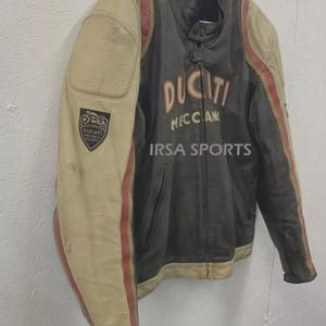May include: A vintage Ducati motorcycle jacket in black and cream with red stripes. The jacket features the Ducati logo and the text "MECCAN" on the front. A patch with the Ducati logo is on the left sleeve. The text "IRSA SPORTS" is also visible.