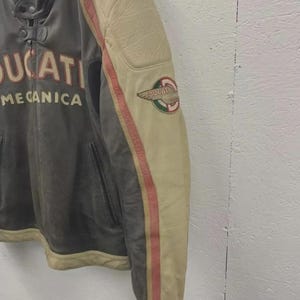May include: A vintage leather motorcycle jacket with the word "DUCATI" and "MECCANICA" printed on the front. The jacket features a cream-colored sleeve with red stripes and a Ducati logo patch. The jacket is a mix of gray and cream colors.