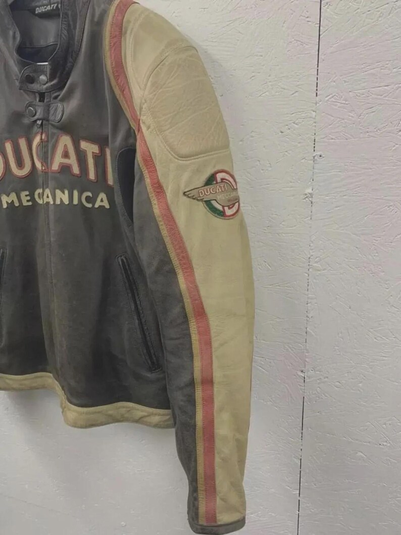 May include: A vintage leather motorcycle jacket with the word "DUCATI" and "MECCANICA" printed on the front. The jacket features a cream-colored sleeve with red stripes and a Ducati logo patch. The jacket is a mix of gray and cream colors.