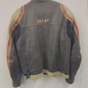 May include: A vintage gray leather motorcycle jacket with the word "DUCATI" across the back. The jacket features tan and red stripes on the sleeves and tan trim along the bottom.