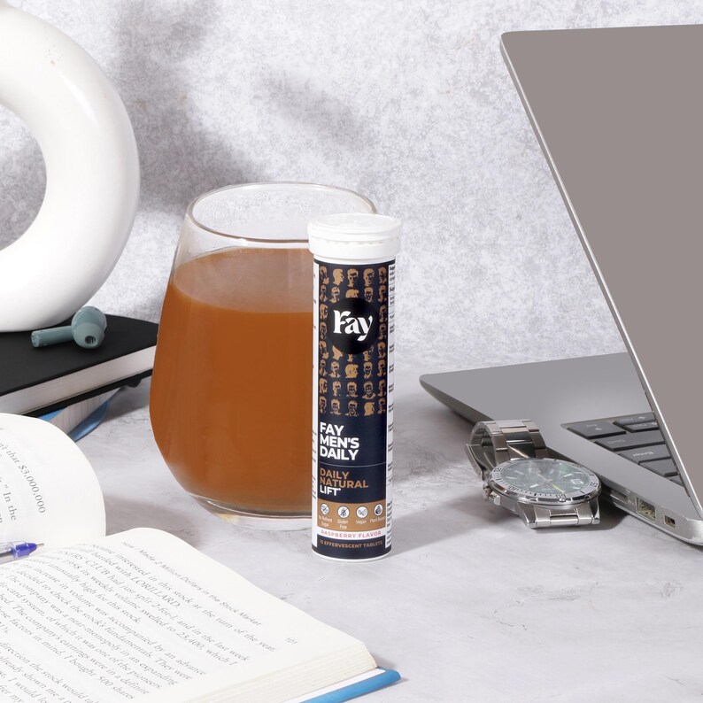 May include: A clear glass of brown liquid sits next to a white and navy blue tube of "Fay Men's Daily Natural Lift" effervescent tablets. A silver wristwatch and a laptop are also on the table.