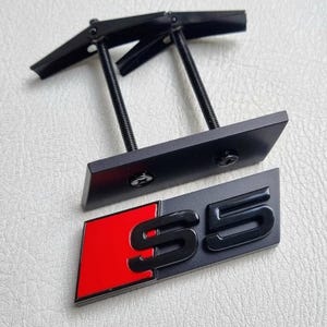 May include: Black and red S5 emblem with mounting hardware. The emblem features a black "S5" logo on a grey background, with a red accent. The mounting hardware is black.