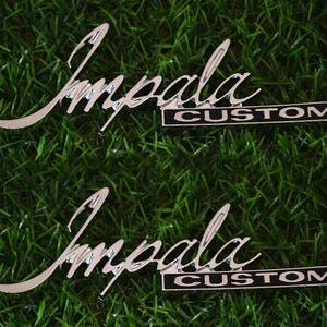 Puede incluir: Dos emblemas Impala Custom plateados sobre un fondo verde y herboso. La palabra "Impala" está en cursiva, y "CUSTOM" está en un diseño rectangular en blanco y negro.
